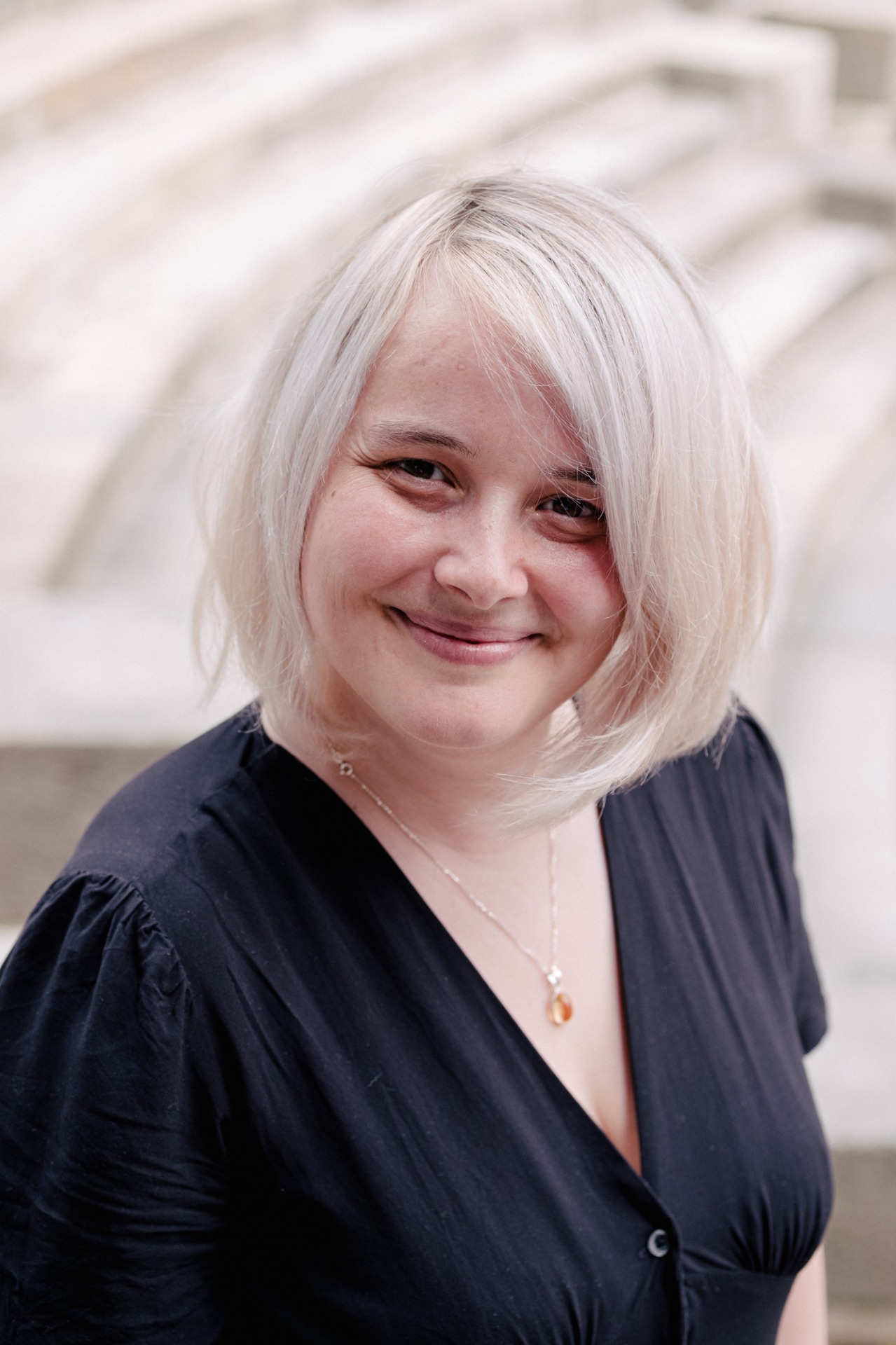 Portrait rapproché d’une femme d'une trentaine d'années aux cheveux blancs, souriante, portant un chemisier noir et un collier fin. Marie BIENVENÜE
